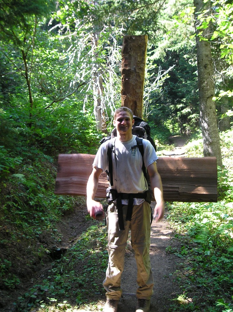 A person carries a backpack with large pieces of wood sticking out of either side along a trail. 