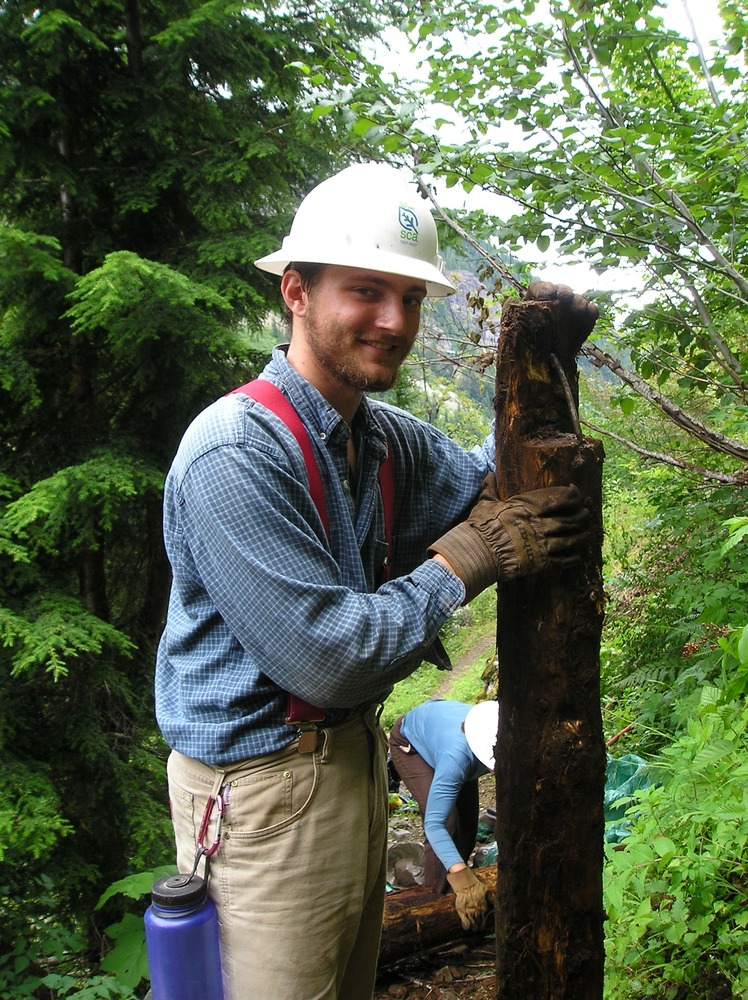 A smiling young man in a white hard hat stands next to an upright post. 