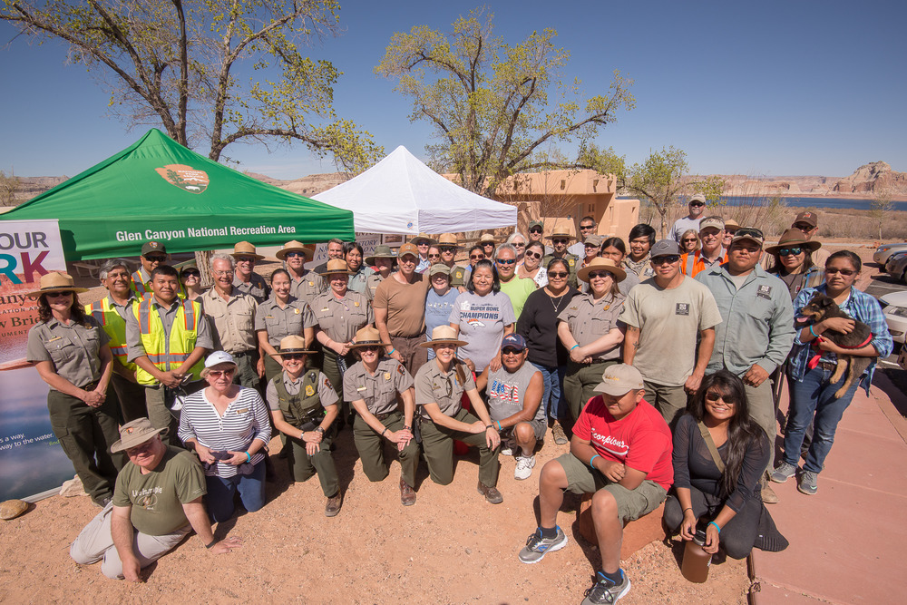 A large group of people, including rangers in uniform. Pop-up tents in the background, Lake Powell in the far background.