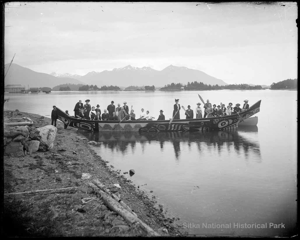 Ceremonial Native dugout canoe filled with 31 people dressed in suits and hats. Pyramid mountains in background. Around 1900.