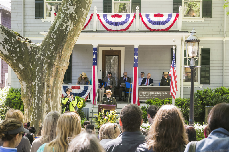 A woman in a hat and green suit speaks at a podium in front of a grey house. 