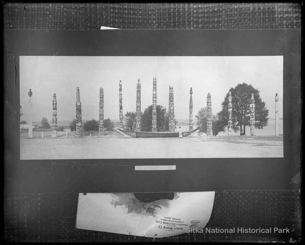 A copy photograph of thirteen totem poles and ceremonial canoe on display.
