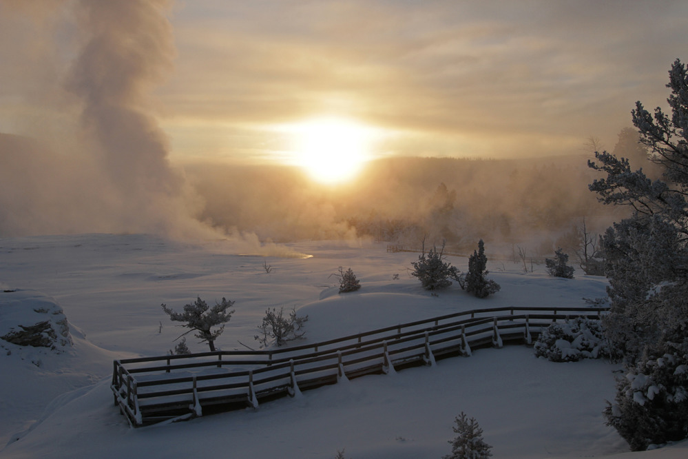 Looking over a boardwalk at snow covered terraces with steam rising on the left and the sun rising in the background.