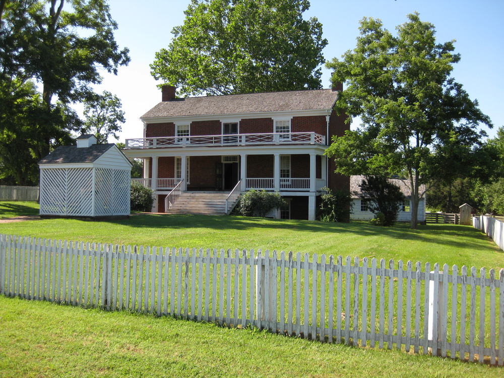 color photo of a red brick house with a full balcony and porch set back on a green lawn and surrounded by a white picket fence