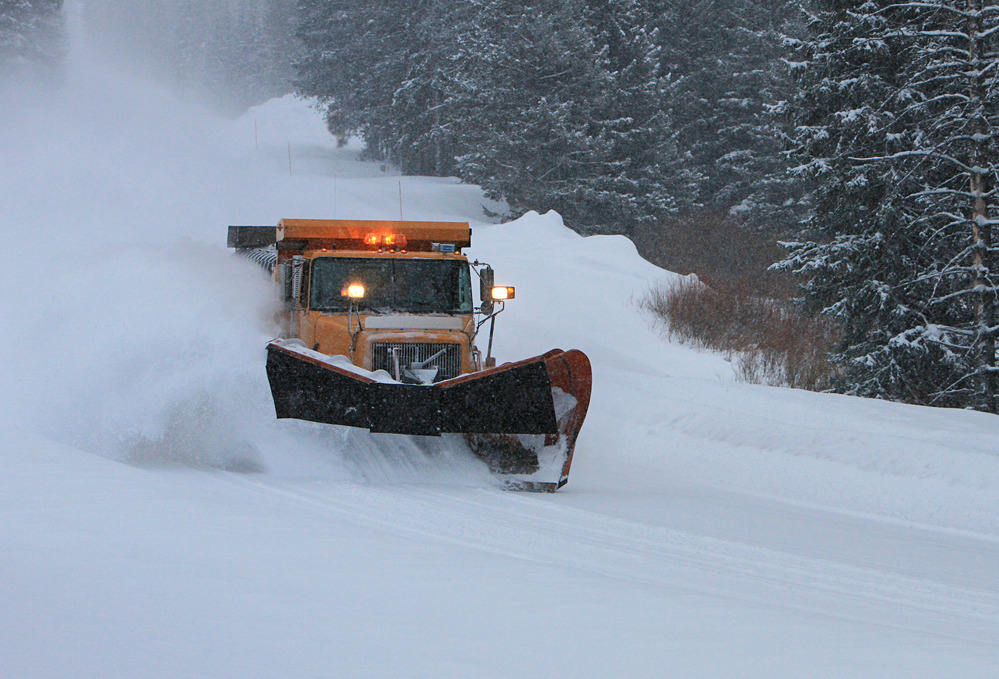 Snow plow on snowy road with forests surrounding it.