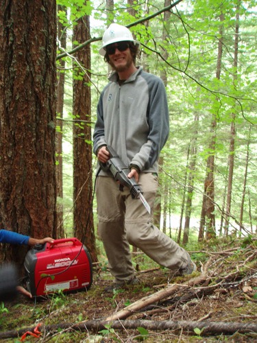 A young man in a hard hat holds a power saw tool connected to a portable charger while standing in a forest. 