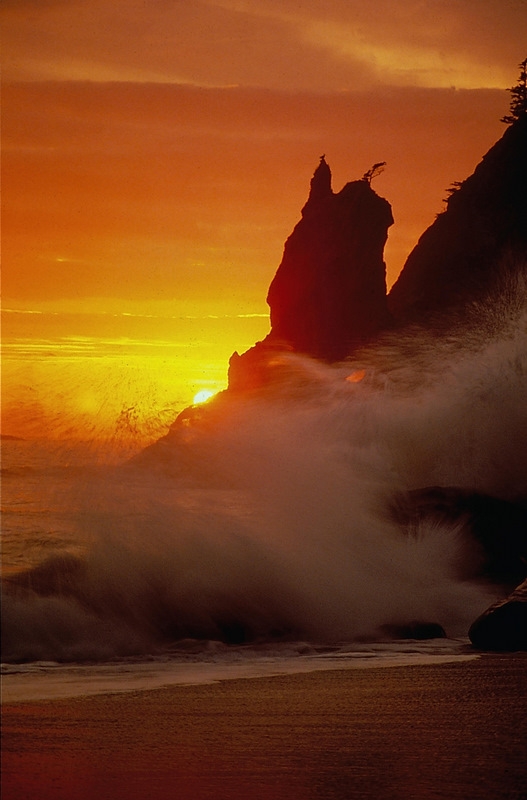 A wave crashes against coastal rock formations at sunset.