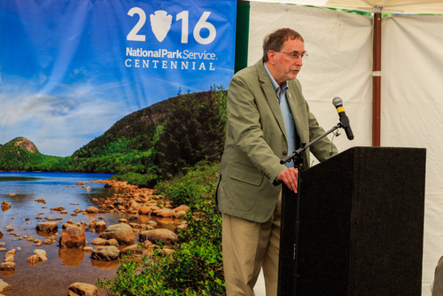 A man stands at a podium speaking in front of a large banner featuring a photo of Jordan Pond and the NPS centennial logo.