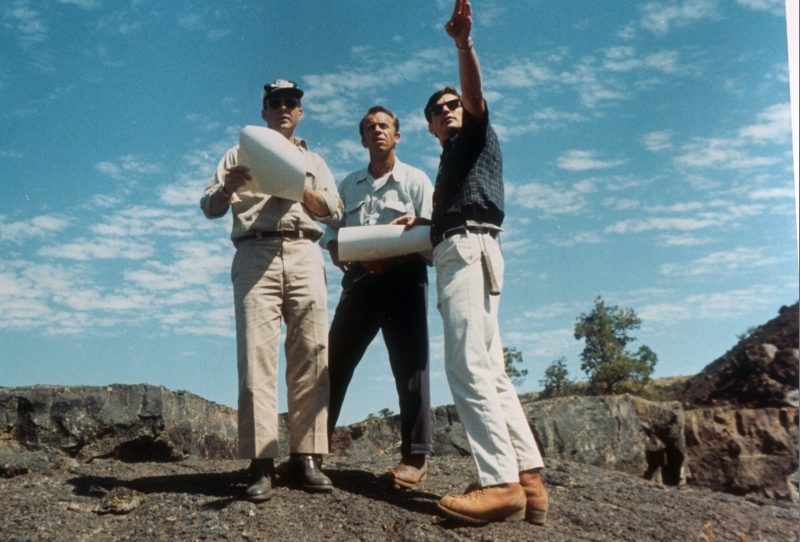 photo of three men holding maps and looking off into the distance, one is pointing