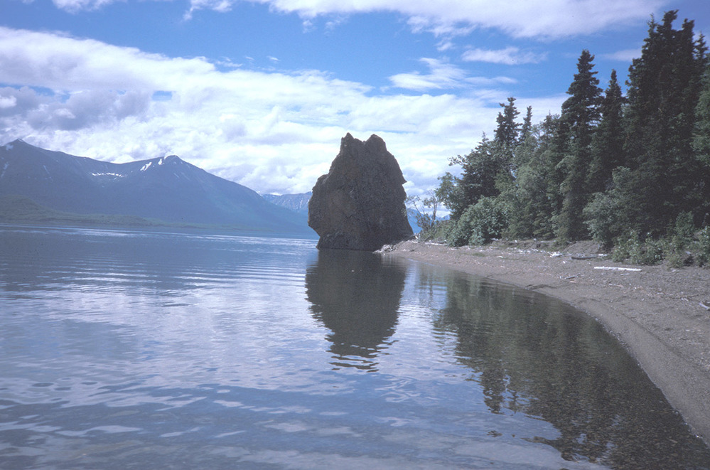 A large rock rises at the end of a curving lakeshore, with mountains in the distance.