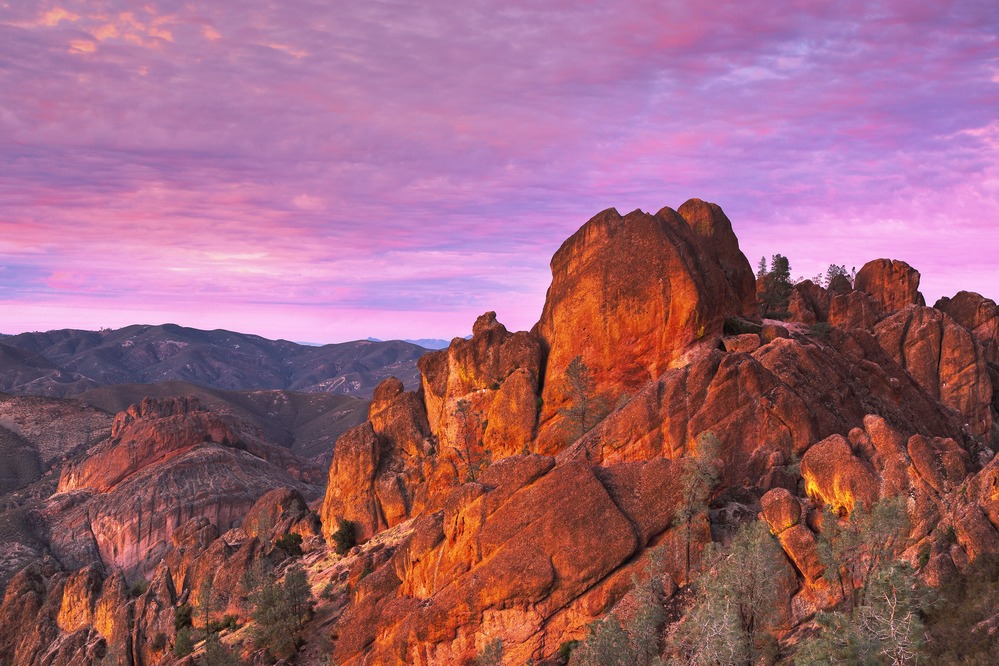 The orange glow of sunset on the rocky spires of the Pinnacles high peaks, with a purple sky at sunset.