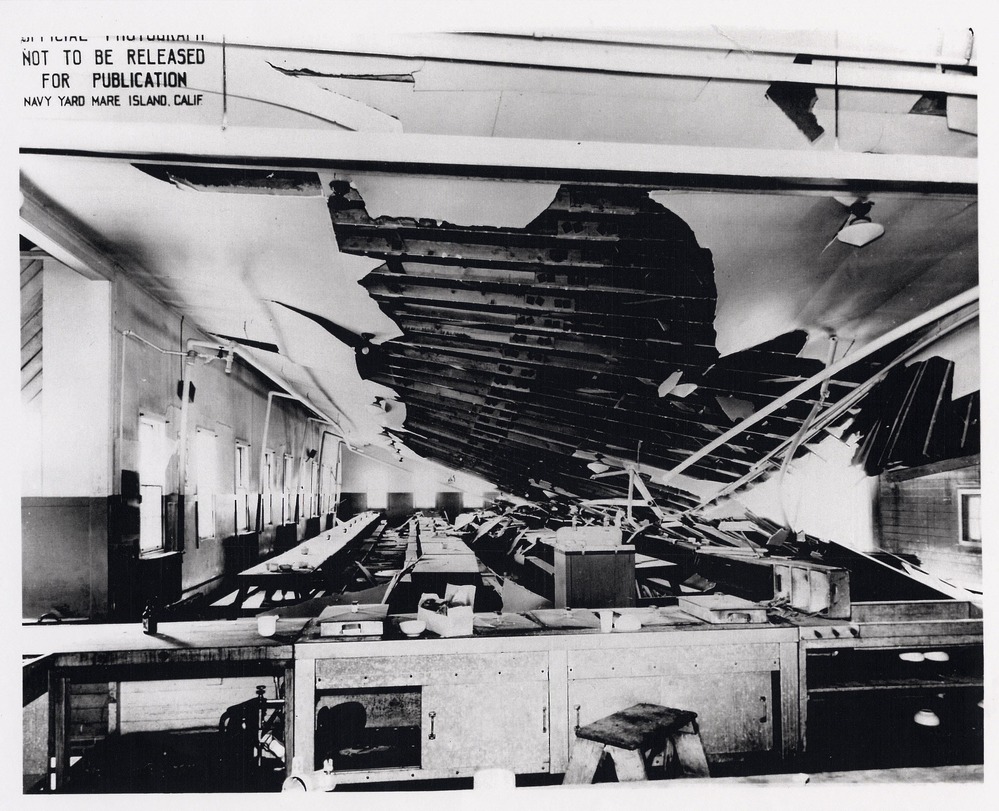 Historic image of the inside of the mess hall after explosion. Ceiling is partially collapsed revealing the roof  beams. Large cafeteria like room with tables is in disarray.