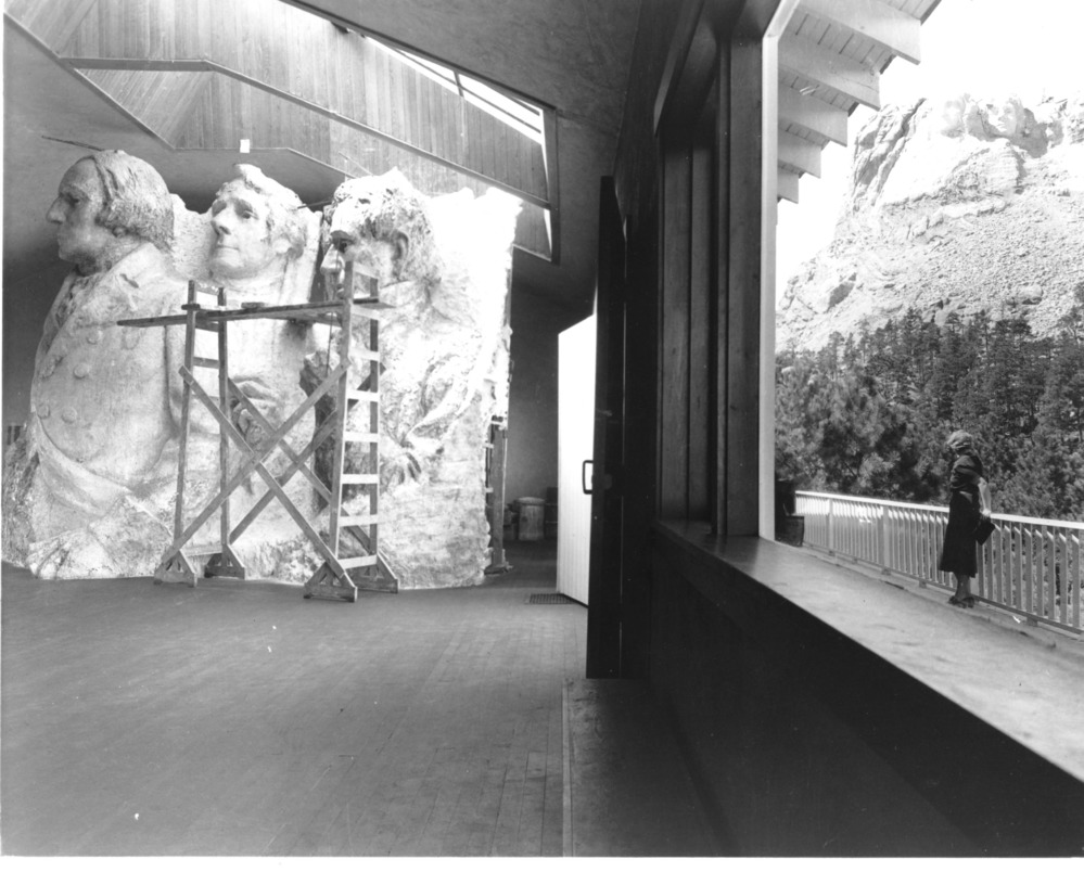 View of the interior of the first on-site sculptor's studio with a large window to the right.  A woman wearing a dark dress is standing outside leaning against a railing with Mount Rushmore in the background.  The left side of the image show the scale model made of plaster with a scaffold in front of the model.