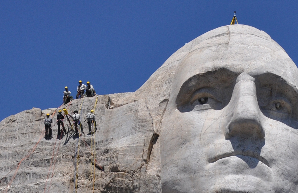 Mount Rushmore National Memorial technical ropes team members work with a custom made tripod that will be used in the scanning of the sculpture.  Four team members are suspended by ropes to the left of George Washington's carved face while three team members assist from above.