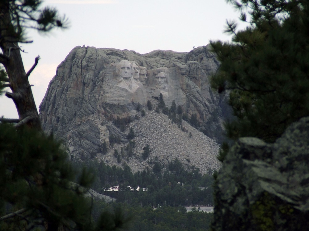 A distant Mount Rushmore framed by ponderosa pine trees and rocks viewed from Iron Mountain Road on an overcast day.