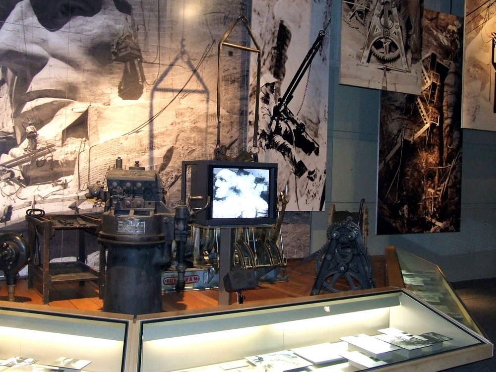 Photo of the Exhibit Hall showing equipment used by blacksmiths (left side of image) and a winch (right side of image) used during the carving of Mount Rushmore.  Numerous historic photos of workers and equipment hang in the background.