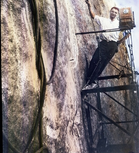 Charles D'Emery, a white male wearing dark pants and a white shirt, poses with his camera on metal scaffolding attached to a vertical granite wall.