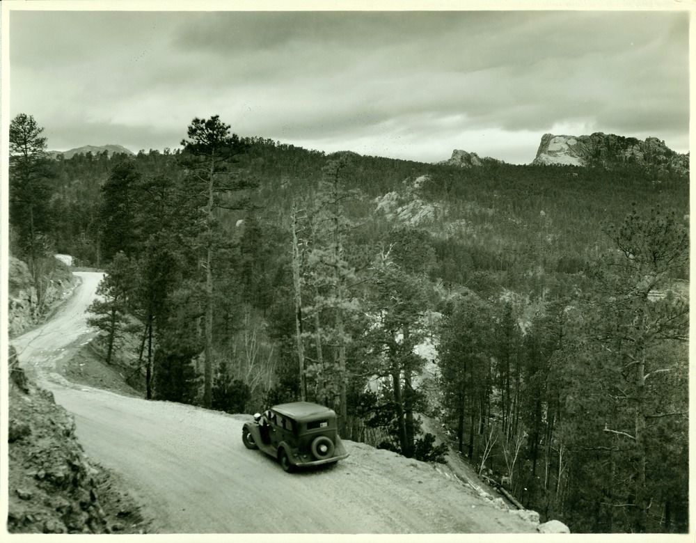 Black and white image of Mount Rushmore from Iron Mountain Road.  Mount Rushmore looks to be about 3 or 4 miles away in the upper right.  The Iron Mountain Road winds its way from the bottom center to the middle right with a dark-colored car sitting on the road near the bottom center of the image.
