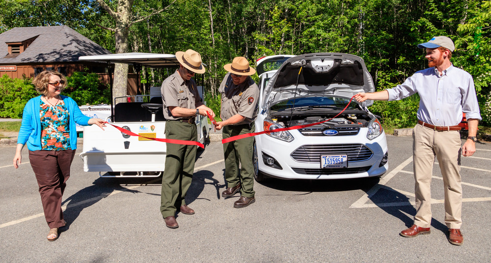 Two rangers cut a ribbon while two others hold the ends during a ceremony welcoming new greener vehicles to the park fleet.