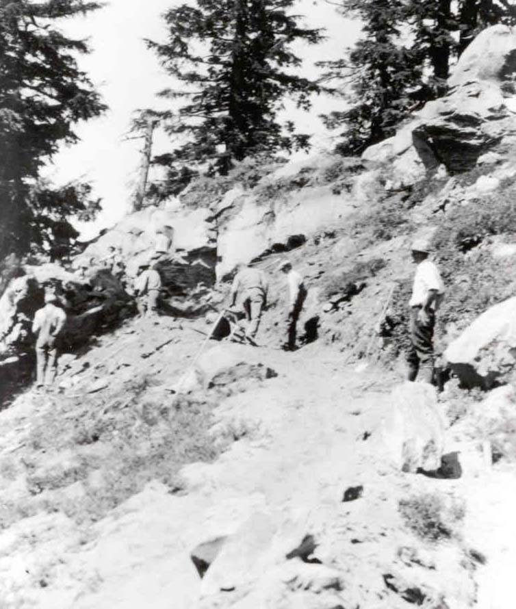 A crew of men with shovels and tools are spaced along a trail that follows the side of a steep, rocky slope.