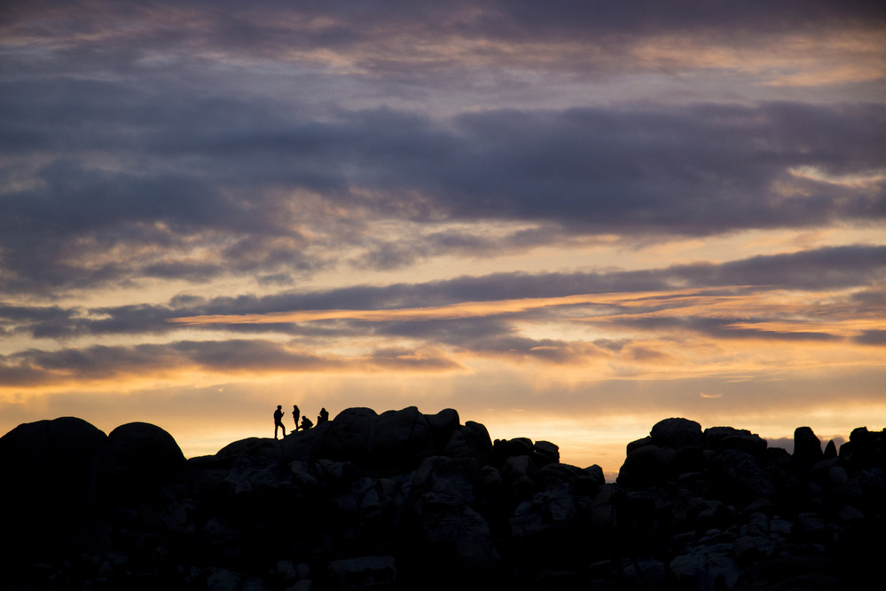 visitors on rounded rocks in the distance are silhouetted against a colorful sunset sky