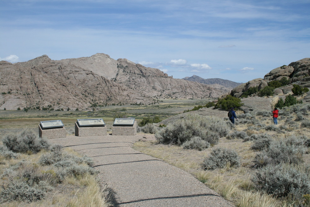 Two visitors walk on a path facing a large rock buttress. A paved trail leads to three wayside exhibits.