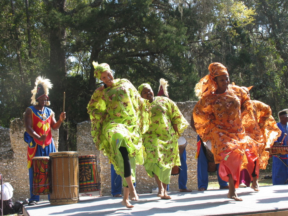 dancers in vibrant colored costumes perform in front of tabby slave cabins