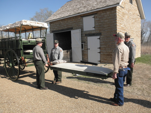 two park rangers and a man load a medical strecther into the back of the green covered wagon