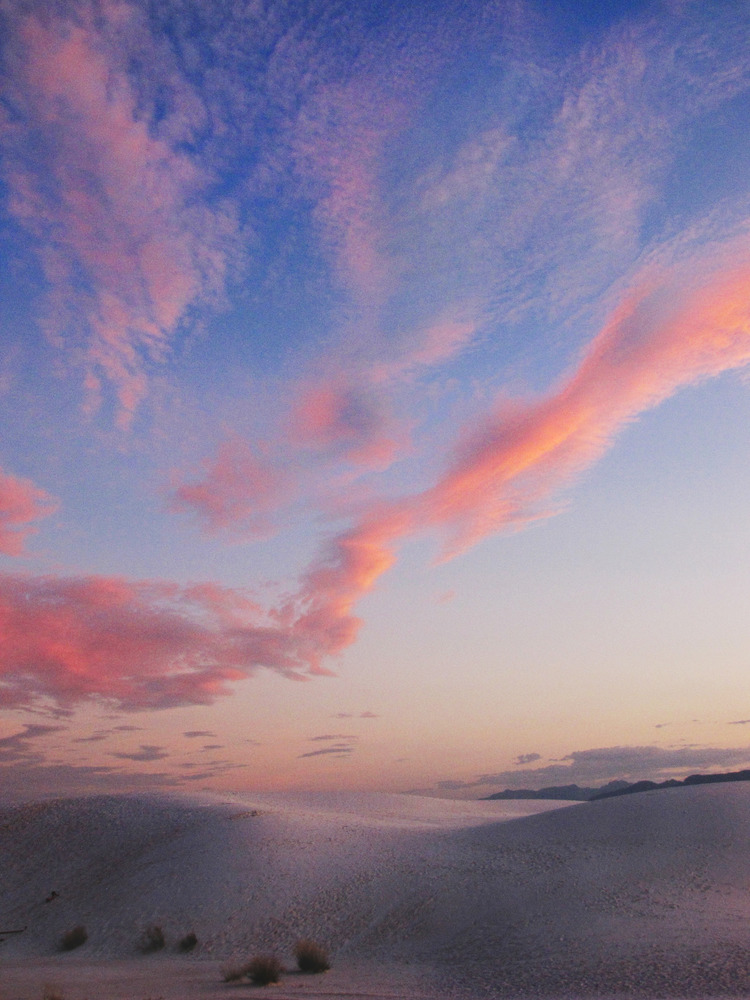 Lines of coral, pink, and orange clouds in a deep blue sky over the dune field.
