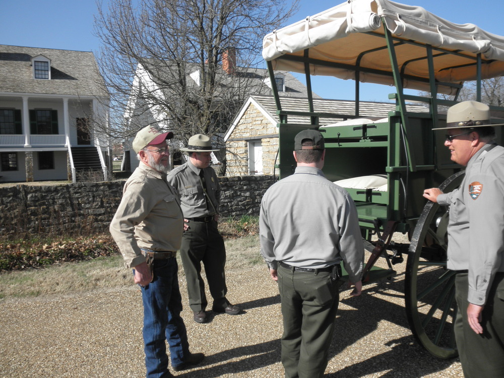 Three park rangers and another man gather around rear of green covered wagon.
