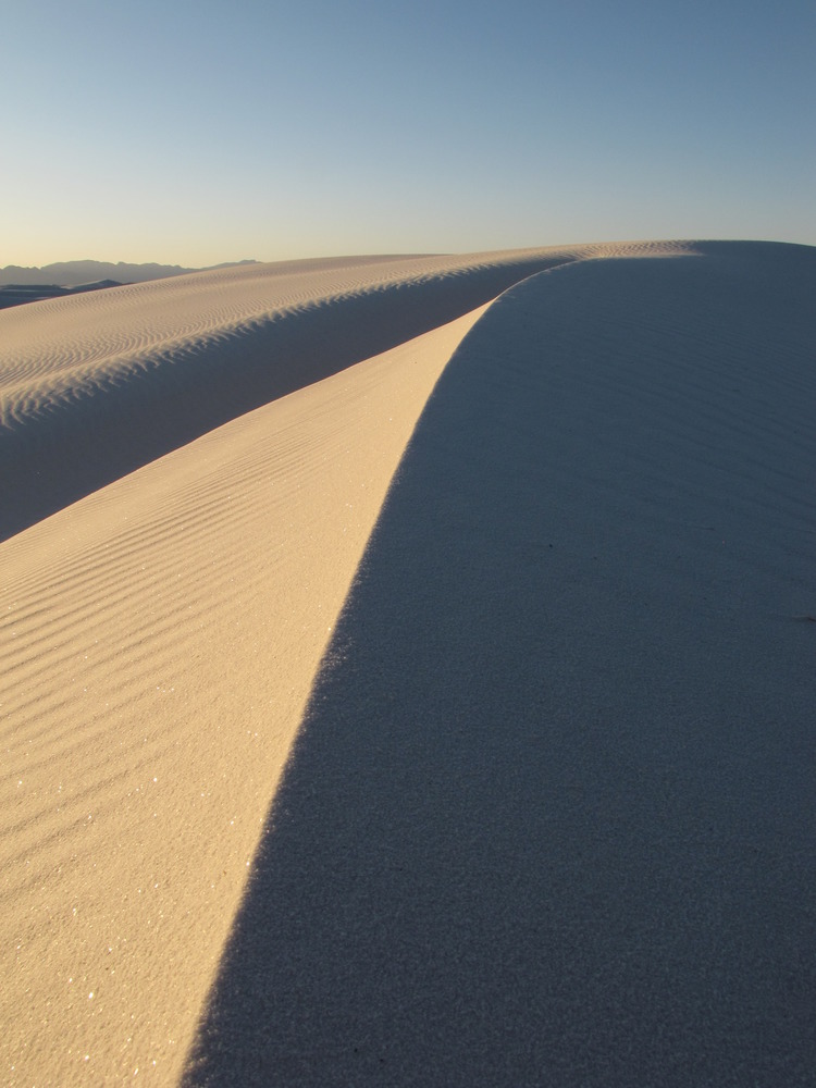 The light of the sun perfectly illuminates one side of a sand dune, while the opposite side is dark. A sharp, straight line at the highest point of the dune separates the two sides.