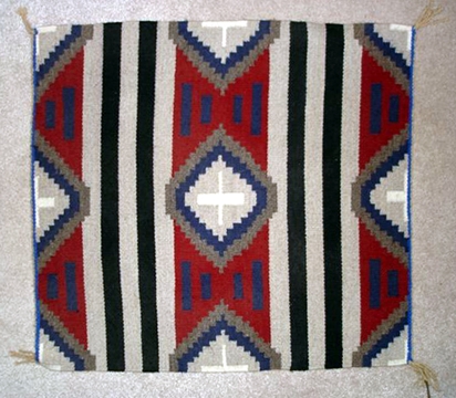 Navajo Rug, Third Phase Chief design. Colors are primarilly red, blue, white, and gray. Often square. Triangles in the corners and a diamond in the center. Striped without a border.