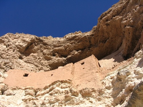 Looking up at a large cliff dwelling inside a cave entrance with several small windows and peep holes. The lower half of the dwelling is a darker reddish color and the upper half, which is barely visible, is a lighter tan color that matches the cliff face. 