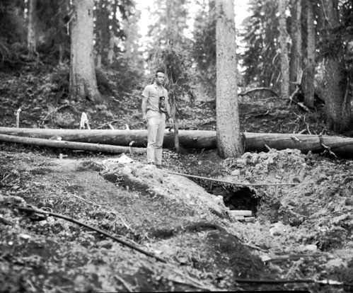 Man with camera stands next to water supply hose and pipes (middle ground and foreground) in forest. Record of water exploration.
