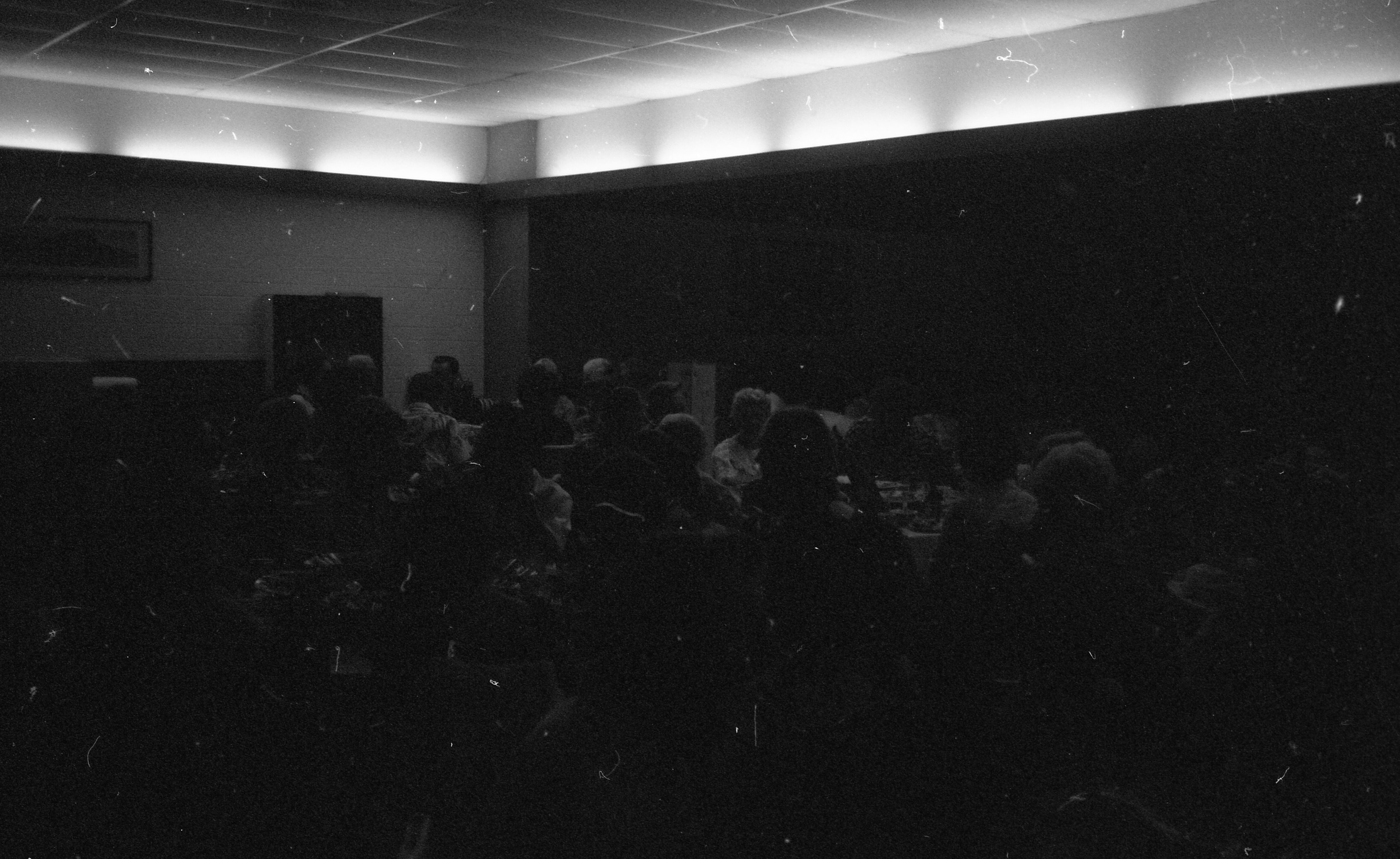 A very dark black and white image of a large group of people sitting around tables in a large room. The tables are circular with people sitting around them. There are glimpses of dishes and glasses on the tables. At the top of the image there is a drop-down ceiling. There are lights along the top of two walls behind the tables that illuminate the top fourth of the walls.
