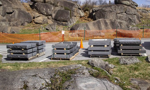 There are several stone slabs set into wooden storage bundles placed along a parking lot. The area is fenced off with orange construction fencing. In the foreground and background are large boulder formations.