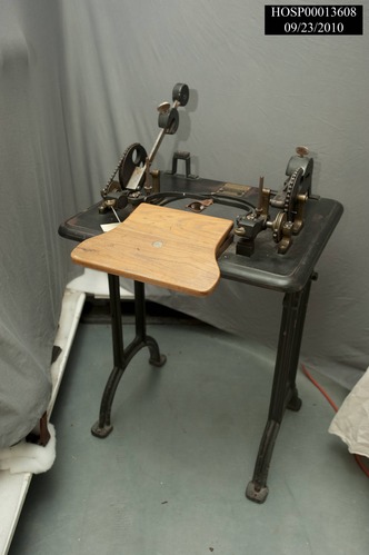 "Zander Type A-12 Adjustable Weight Finger Joint Unit" is a cast-iron device that resembles a typing table. The center has a large U-shaped opening with a wooden hour-glass shaped armrest attached to the front of it, leaving half the armrest hanging over the edge of the table and half of the opening unblocked. A slender curved bar hangs over the space with a finger stall in the center of it. Its ends are attached to weights and gears on each end of the table top. The entire device is supported by two pairs of parallel iron bars, each attached to a cast-iron arc that forms a flat foot at each end that can be bolted securely to the floor. To use this device, the user rests the forearm and hand on the wooden surfaces and places a finger into the leather finger stall, which is set into a two-pronged iron fork. The finger can then be either flexed using the weighted lever on the right or extended using the one on the left, and the respective levers are weighted differently.