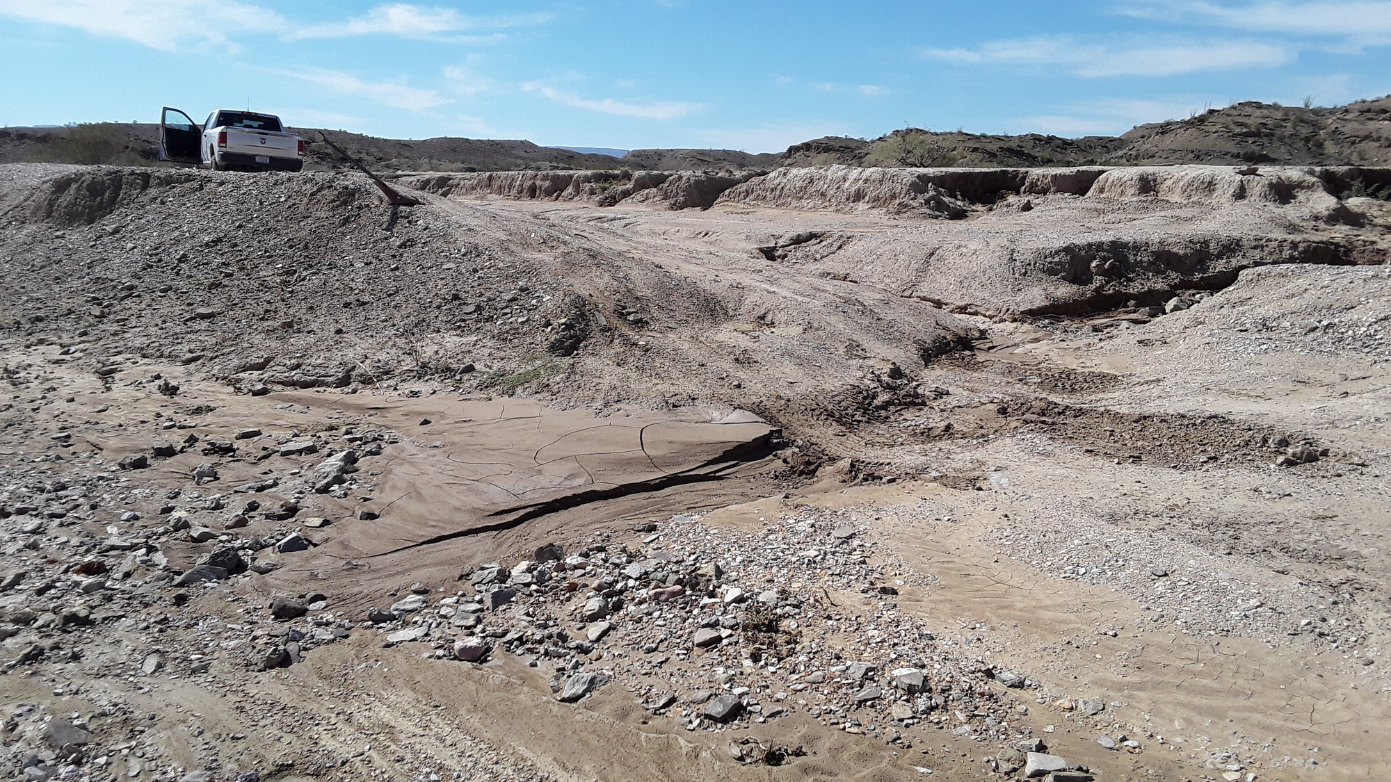 Tire tracks are visible crossing a muddy, gravelly washout on an unpaved road. The tracks lead out of a drainage, through wet mud, and up a slope banked steeply to the right. A truck is parked at the top of this bank.