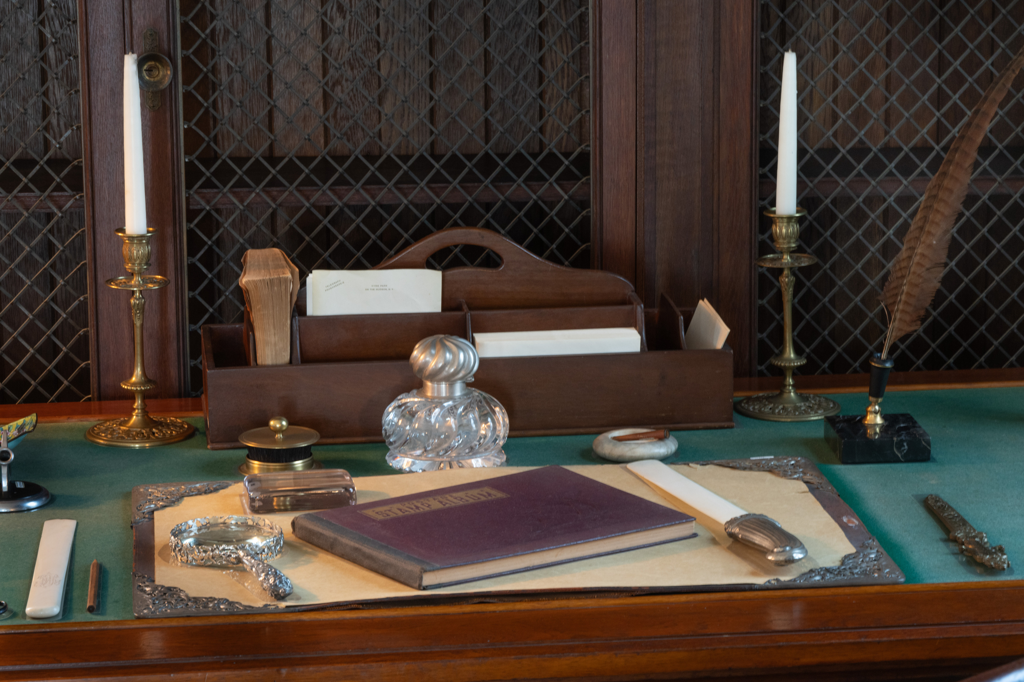 A wood desk with green baize cloth is furnished with bronze candlesticks, a wood letter box, writing quill in stand, magnifying glass and stamp album among other desk accessories.