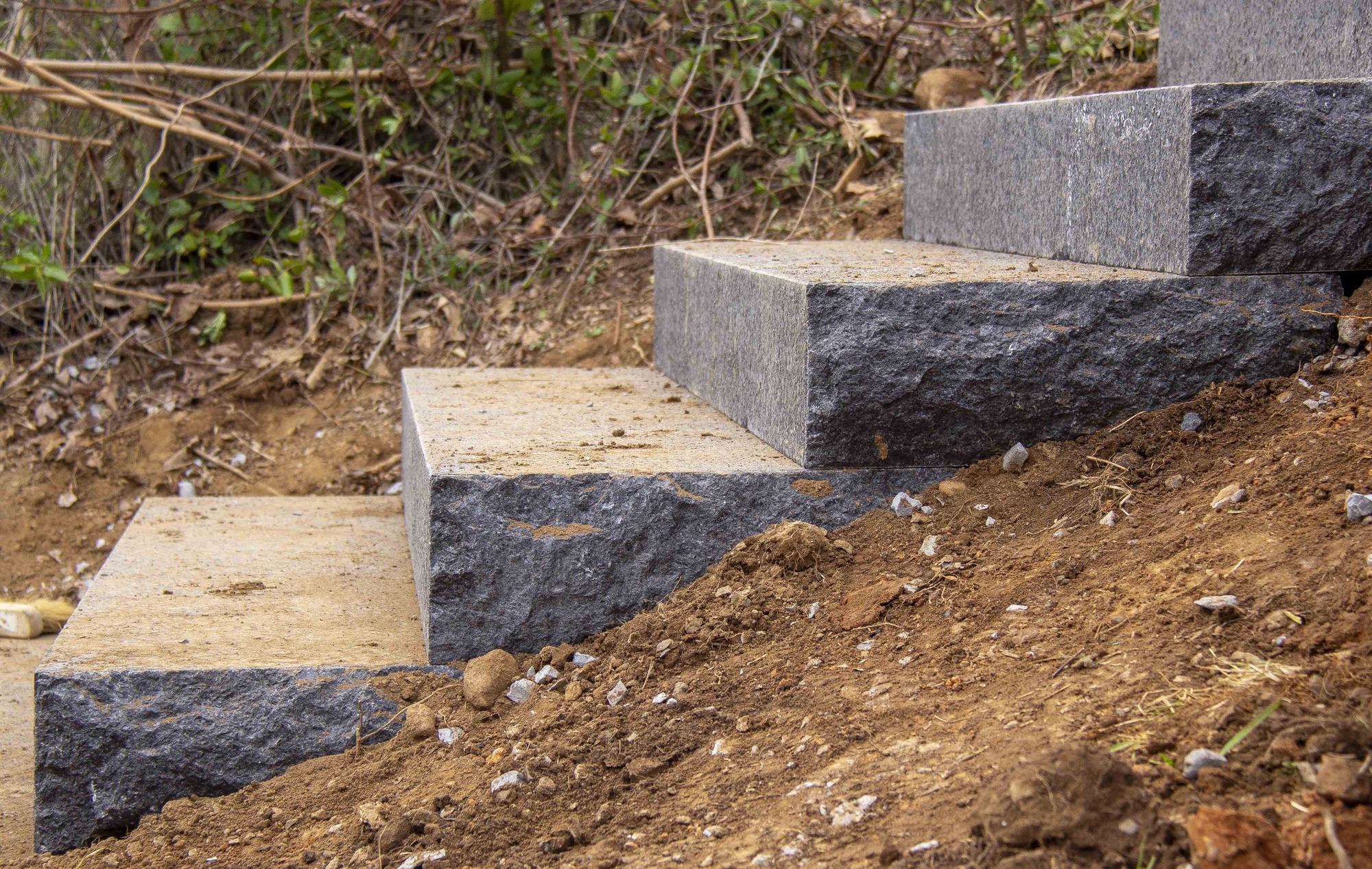 This is a side-view close-up stone of five new stone stairs in the side of a hill. The soil in the foreground is fresh and brown. There is a new layer of dirt on the steps.