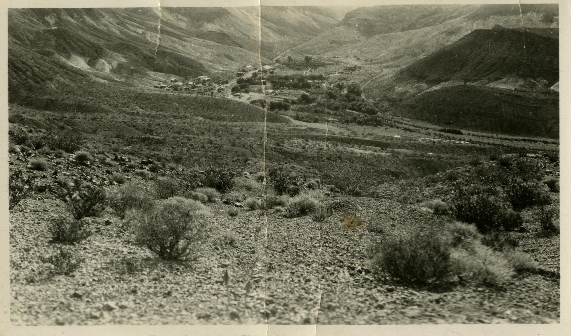 This is an historic black and white photograph from the Scotty's Castle Historic Photograph Collection, Death Valley National Park of building complex settled in wash of desert canyon. Desert gravel, rubble, and shrubs in fore and middle ground. Road and fence line leads into canyon from right, through complex, and out middle background.