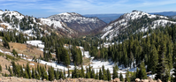 A panoramic photo of a volcanic landscape with patchy snow and numerous conifers.