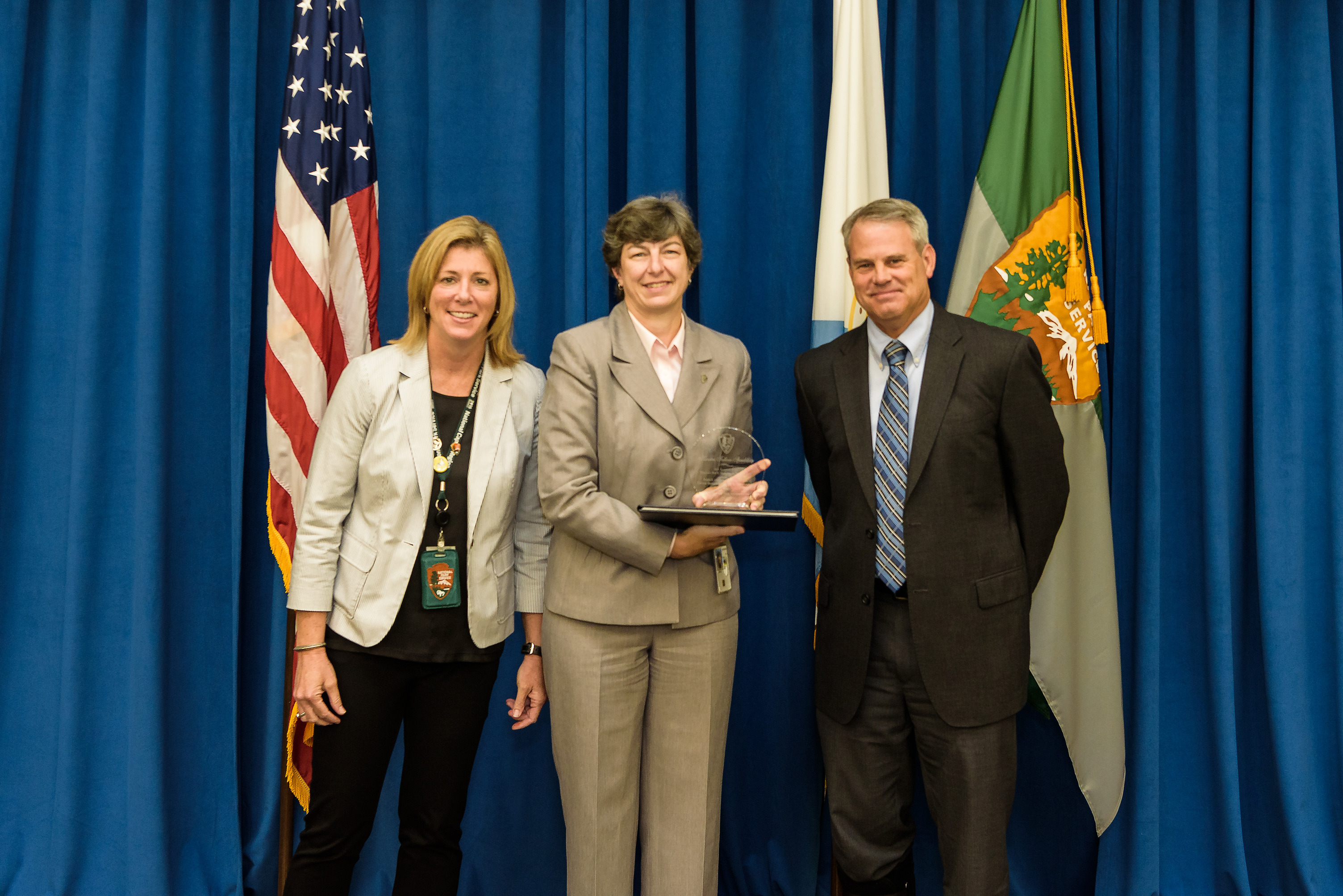 a woman in a tan business suit holds a glass statue and stands between a man and a woman in business suits. The background is a blue curtain, an American flag, and a National Park Service flag.