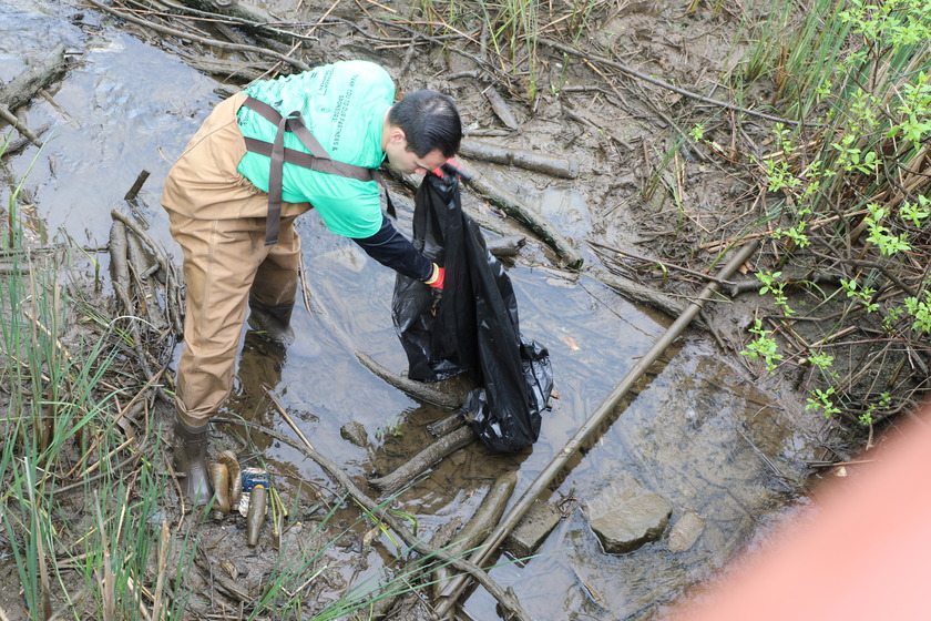 A person wearing a light green t-shirt, tan waders, and work boots is bent over in a muddy stream, actively putting litter in a large black trash bag. The water is murky, and the banks are muddy with some green vegetation and discarded waste like old cans and sticks.