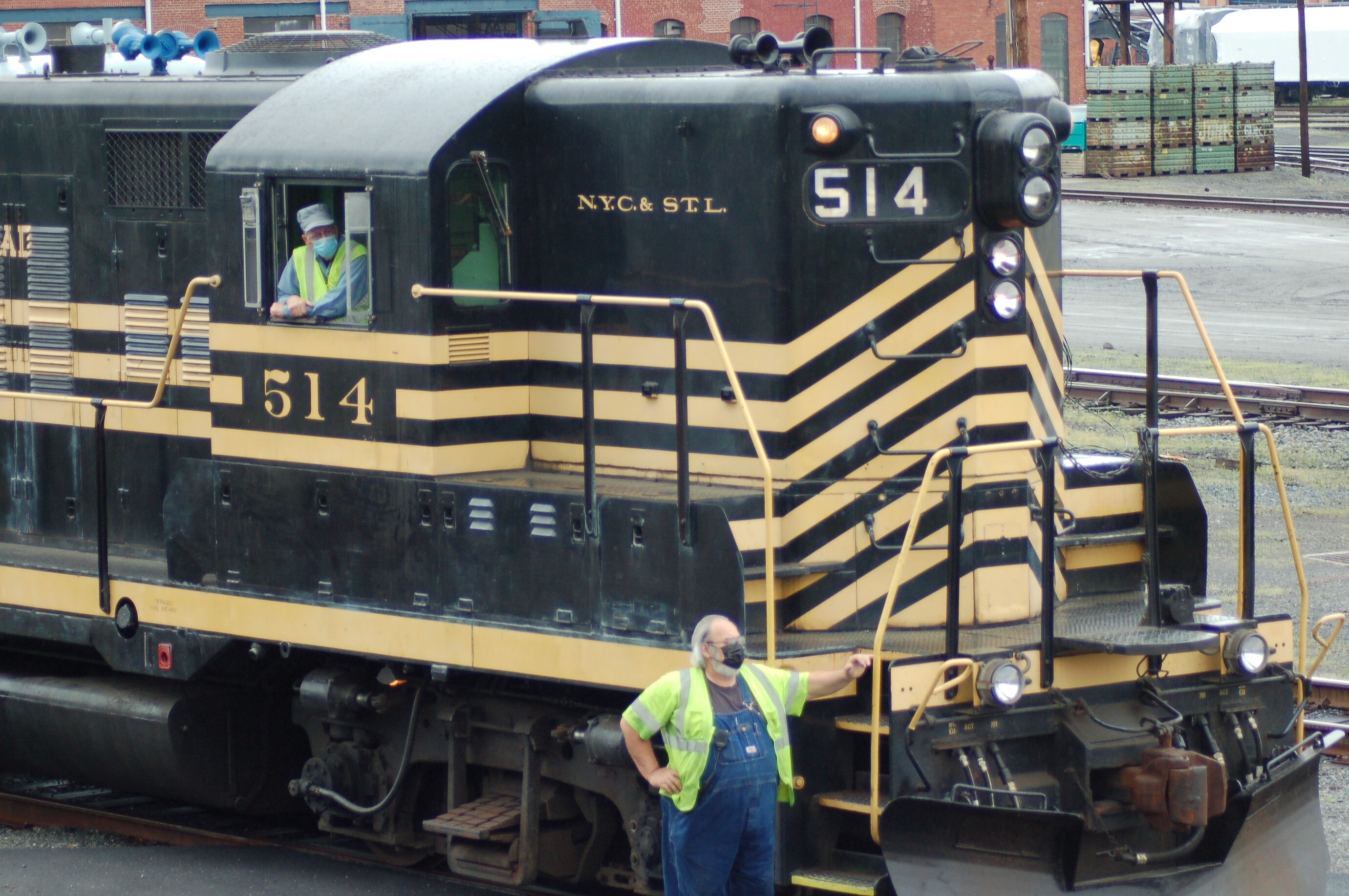 two employees waiting for instruction. one is standing in front of train, the other is up in the cab, peeking out the window