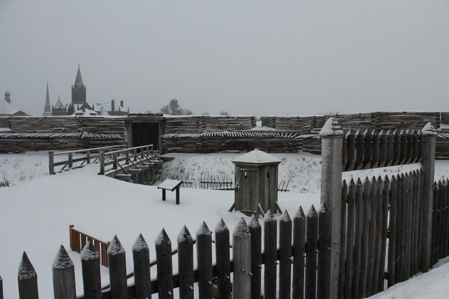 Looking down into an area framed by an wooden picket fence. Inside, a snow covered bridge leads to a large wooden structure behind it.