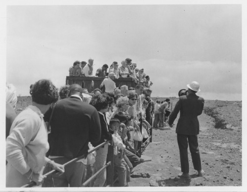 Black and white. A large group of people standing by a fence listening to a man with a bullhorn give a presentation