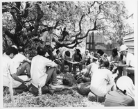 A black and white image of a group of Youth Conservation Corps members and park staff meeting with wildlife ranger, Don Reeser. The group is sitting in a circle in the grass next to a building which is located on the right side of the image. Don Reeser is standing in front of a tree located on the left side of the image. He is wearing a National Park Service uniform including a bottom down short sleeve shirt with a ranger badge and name tag located above the chest pockets, a belt, and pants. He is holding onto a branch of the tree with his left hand. The Youth Conservation Corps members are sitting in a circle around him sitting on top of lava rocks. They are holding a piece of paper in their hands. Some are looking down at the paper in front of them while others are looking at Reeser. In the background a water catchment tank is visible.