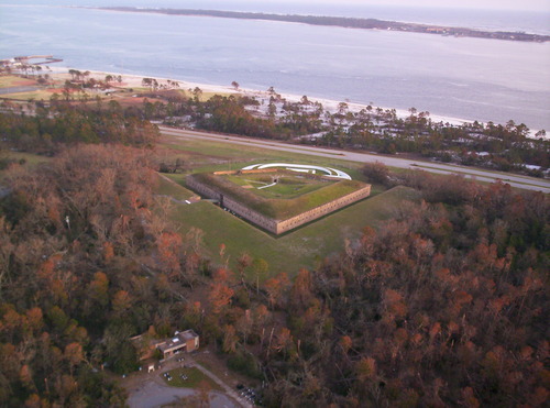An aerial view of a fort built into a coastal island. The beach is visible surrounding one side of the fort: the others are surrounded by trees.