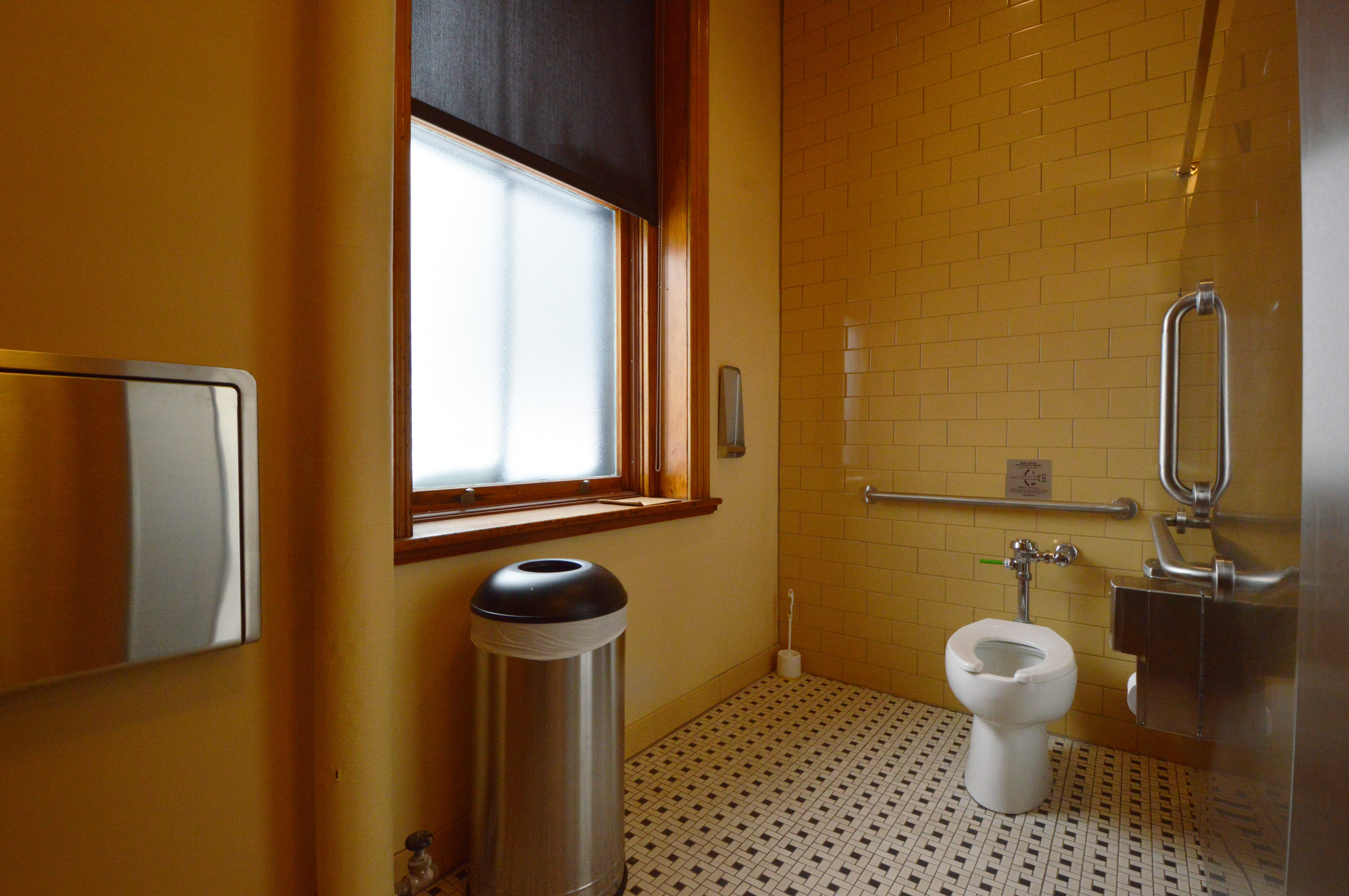 The toilet is in the back right of a stall across from a frosted window. A trashcan is below the windowsill on the left. A portion of the child changing station is seen on the wall at left. 